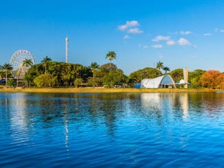 Parque de diversões ao lado de um rio sob céu azul, com roda-gigante, árvores verdes e arquitetura moderna, destacando opções de lazer e turismo na região, com foco em sustentabilidade e meio ambiente.