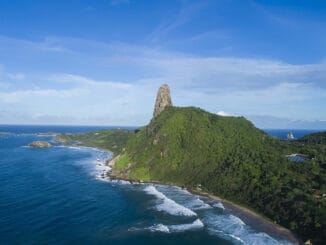Céu azul e mar na costa de uma ilha com vegetação exuberante e formação rochosa distinta, perfeita para turismo ecológico e preservação ambiental no Brasil.