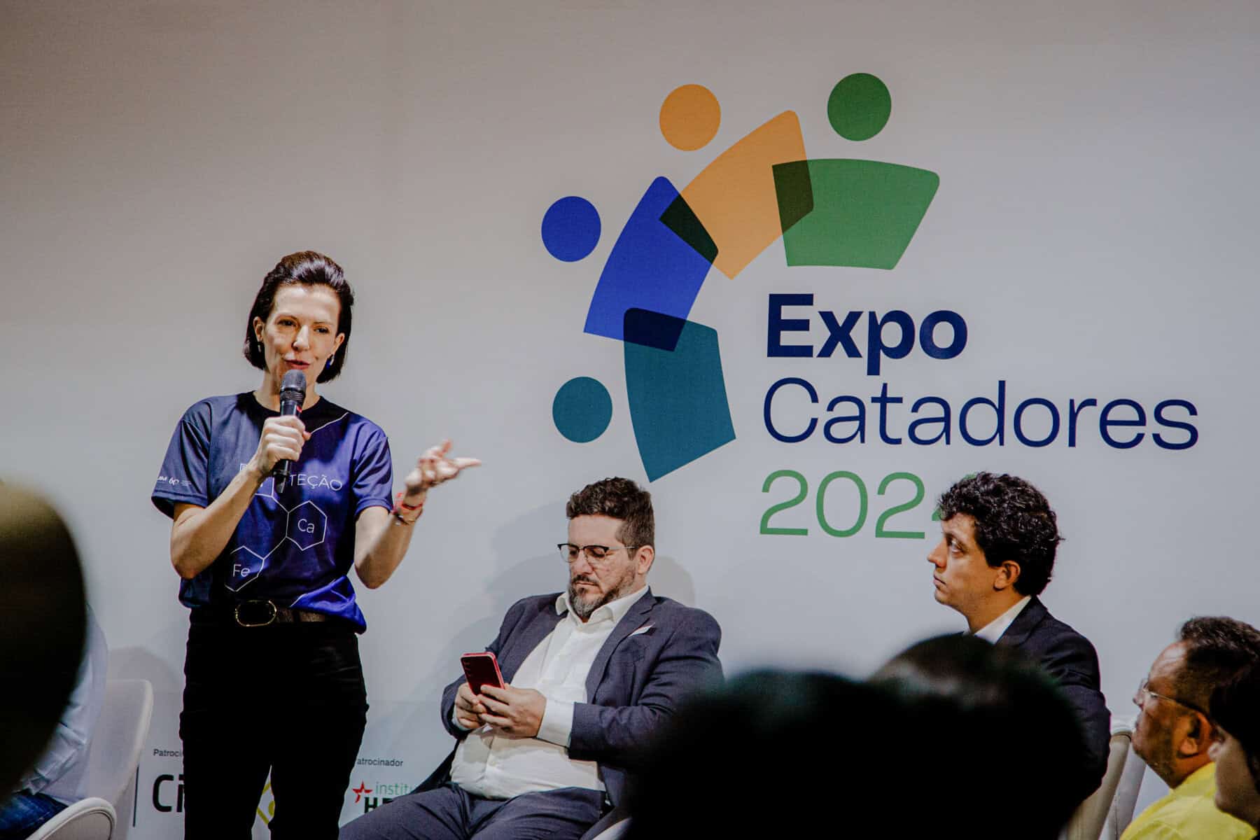 Guarda de mulher apresentando no palco do Expo Catadores 2022, evento sobre resíduos sólidos, reciclagem e sustentabilidade ambiental, com audiência de participantes e painel de debates.