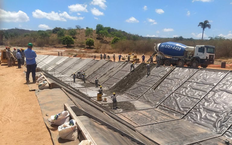 Bone dry cement being poured into a large pre-lined excavation site for construction or environmental remediation. Workers and a cement mixer truck are actively involved in the process.