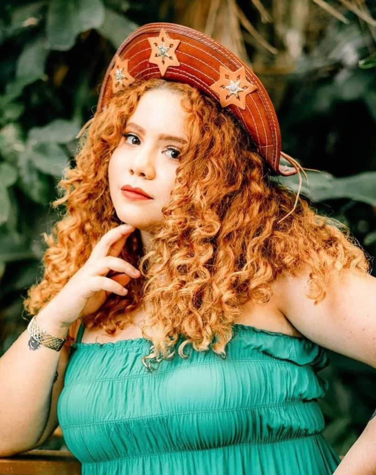 Cenário com mulher de cabelo cacheado ruivo usando chapéu decorado com estrelas de Santa, vestindo vestido verde, com fundo natural de plantas verdes, voltado para sustentabilidade e meio ambiente.