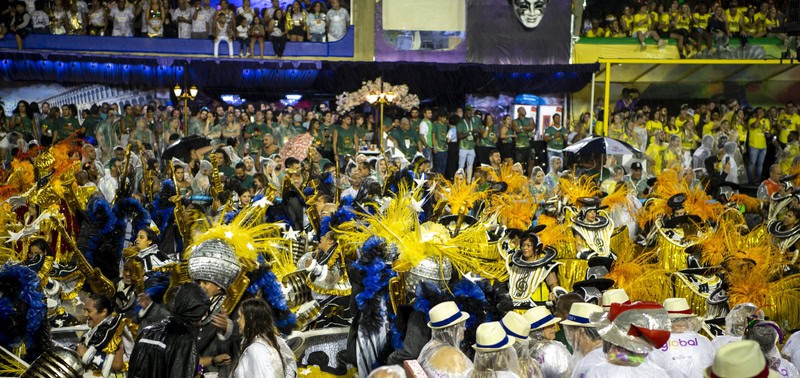 Desfile de carnaval com fantasias coloridas e adereços brilhantes, destaque para o tema ambiental e a celebração cultural no evento. Público diversas pessoas assistindo e participando com alegria.
