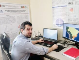 Análise de dados ambientais em escritório, pessoa trabalhando com dois computadores exibindo mapas estratégicos, pesquisa e monitoramento ambiental, foco em sustentabilidade e gestão ambiental.