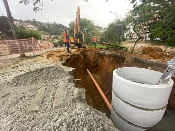 Cabo de fibra óptica sendo instalado em obra de infraestrutura urbana, com escavadeira ao fundo e trabalhadores supervisionando o procedimento, visando melhorias na rede de comunicação. |.