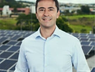 Fotografia de homem sorridente em ambiente com painéis solares ao fundo, promovendo energias renováveis e sustentabilidade ambiental no contexto de notícias ambientais e mercantis.