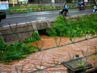 Enchente urbana com água turva atingindo a calçada lado de uma rodovia movimentada com motos e carros. Situação de risco relacionada a mudanças climáticas e gestão ambiental.