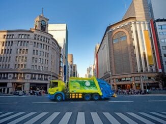 Caminhão de coleta de lixo na rua de uma cidade grande, com edifícios comerciais ao fundo, promovendo limpeza urbana e sustentabilidade ambiental.