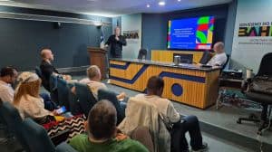 Profissionais participando de palestra em ambiente corporativo, com tela de apresentação digital ao fundo, durante evento de sustentabilidade e negócios ambientais.