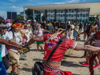 Homens e mulheres indígenas realizando dança tradicional durante evento cultural ao ar livre com estrutura moderna ao fundo, celebrando cultura e sustentabilidade no Brasil.