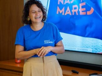 Pessoa sorridente em entrevista ou apresentação, usando camiseta da Ambiental Mercantil, com tela ao fundo exibindo a marca e um símbolo do mar, representando o compromisso com oceanos e sustentabilidade.