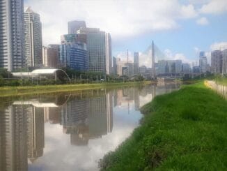Modernidade e natureza no coração de São Paulo com arranha-céus refletidos em um rio sob céu claro.