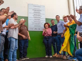 Confraternização de membros da Ambiental Mercantil durante evento de inauguração de uma nova sede ou projeto ambiental. Pessoas sorridentes, aplausos e orgulho em um ambiente interno com parede verde.
