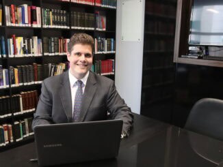 Homem de negócios em escritório, usando terno e gravata, sentado em mesa de reunião com laptop, biblioteca ao fundo, ambiente profissional para temas de sustentabilidade, direito ambiental e negócios verdes.