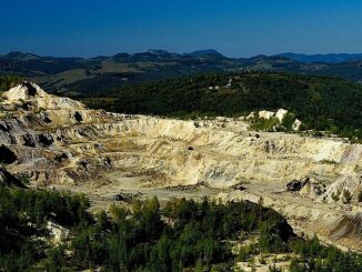 Mineração a céu aberto em área de vegetação e montanhas ao fundo, com céu azul claro.