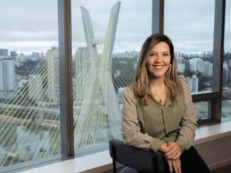 Edifício moderno com vista urbana ao fundo, mulher sorridente sentada próximo à janela de escritório com paisagem de cidade e ponte suspensa ao fundo.