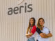 Vestidas com camisetas brancas e vermelhas, duas jovens sorridentes posam de braços cruzados em frente à fachada da Aeris, empresa de soluções ambientais, promovendo sustentabilidade e inovação ambiental.