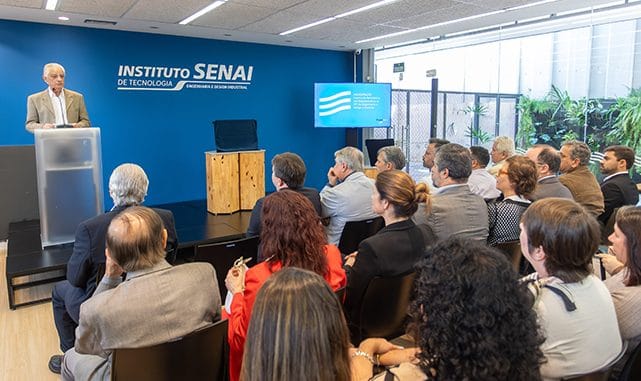 Palestrante apresentando em evento do Instituto SENAI com audiência de profissionais em ambiente moderno de sala de conferências.