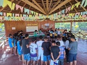 Grupo de estudantes participando de atividade ambiental ao ar livre em espaço com cobertura de madeira e decoração colorida.