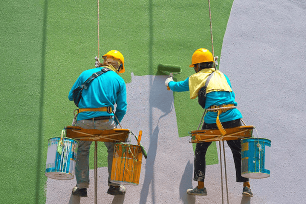 Two painters applying green and gray paint to a wall from scaffolding.