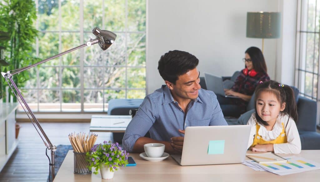 A man is working from home on his laptop at a desk with a young girl beside him, while a woman enjoys home comfort using her laptop on the sofa in the background.