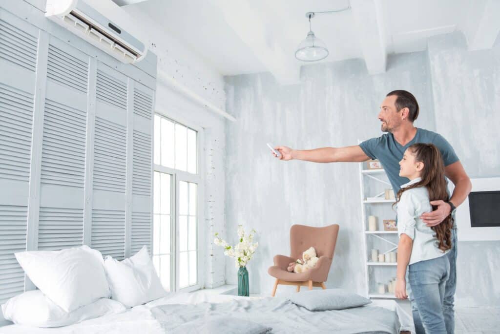 A man uses a remote control to operate a mini split cooling system while standing with a young girl in a bright bedroom.
