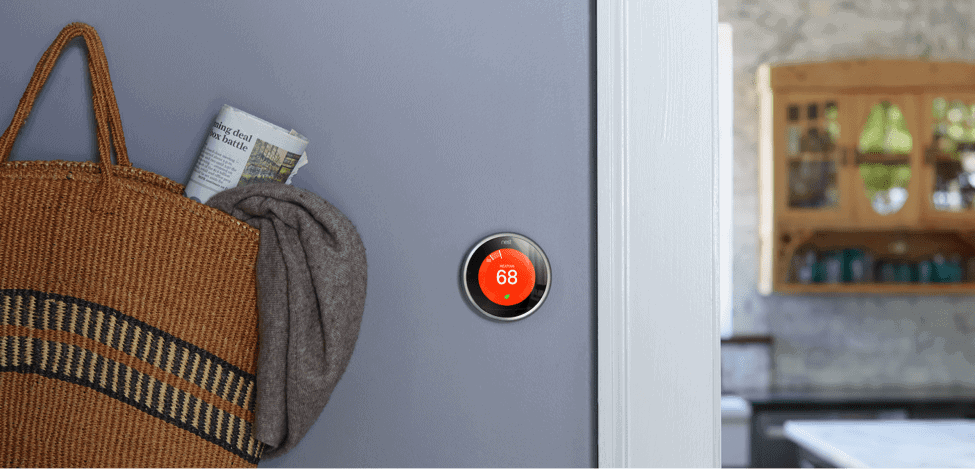 A Nest smart thermostat set to 68 degrees is mounted on a gray wall next to a hanging woven bag with a newspaper and cloth, with a kitchen visible in the background. Enjoy the benefits of modern convenience and energy efficiency.