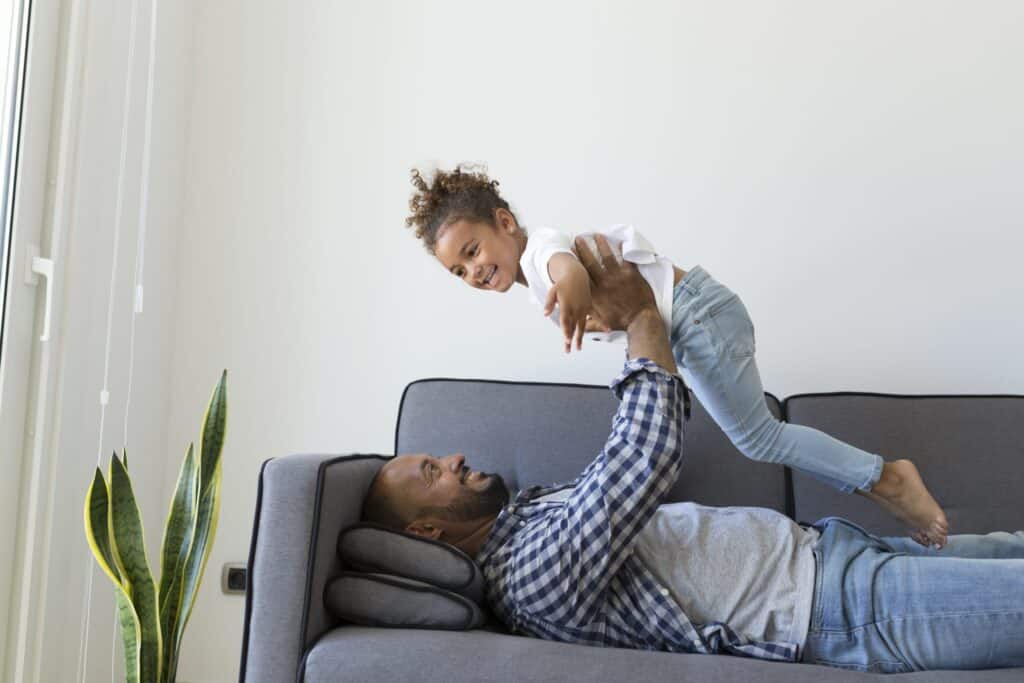 An adult lying on a couch lifts a smiling child into the air; both appear relaxed in a bright living room with a plant nearby, enjoying comfort from an efficient home heating and cooling system.
