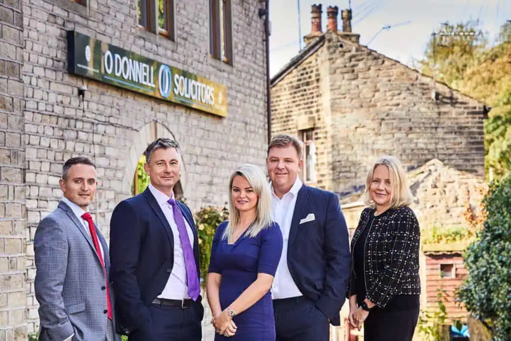 A group photo of five professionally dressed individuals standing in front of a stone building with a sign that reads "O'Donnell Solicitors." The individuals appear confident and approachable, reflecting the firm's professional and community-focused ethos. The setting includes traditional architecture and greenery, emphasizing a local, welcoming atmosphere.