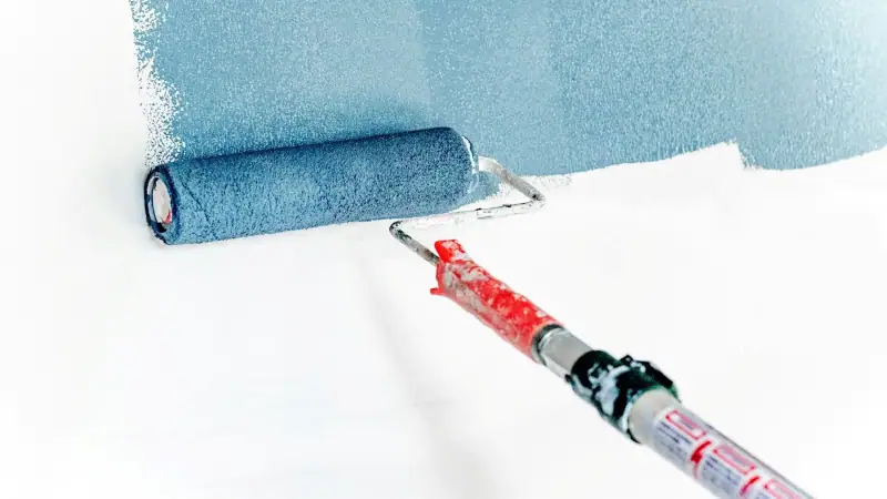 A paint roller laying down light blue paint onto a white wall.