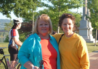 Two women smiling at a park trail event, with a cyclist in the background.