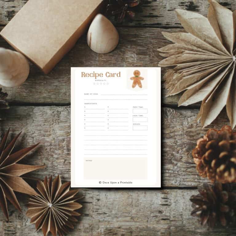 Gingerbread man watercolor graphic illustrated recipe card sitting on a wooden surface with paper snowflakes and pinecones.