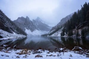 Snow-covered mountains and evergreen trees reflected in a calm lake under a misty sky.