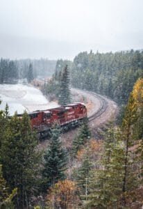 Red Canadian Pacific train on curved railway tracks in snowy forest near a frozen river.