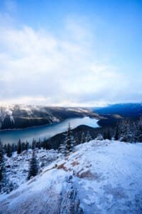 Snowy mountain view with a winding lake and dense evergreen forests under a partly cloudy sky.