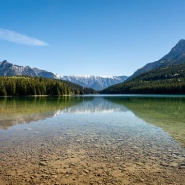 Clear lake with rocky bottom bordered by evergreen trees and snow-capped mountains under blue sky