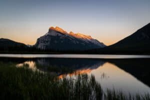 Mountain peak glowing orange at sunrise reflected in calm lake with grassy shore.