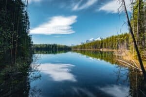 Calm lake reflecting evergreen forest, snow-capped mountains, and clouds under a clear blue sky.