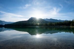 Sun rising over a mountain with forest reflected in the calm clear lake under a blue sky.
