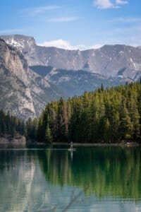 Person paddleboarding on a calm lake surrounded by evergreen trees and rocky mountains.