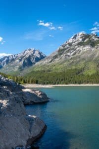 Mountain lake with rocky shoreline, evergreen forest, and clear blue sky with clouds.
