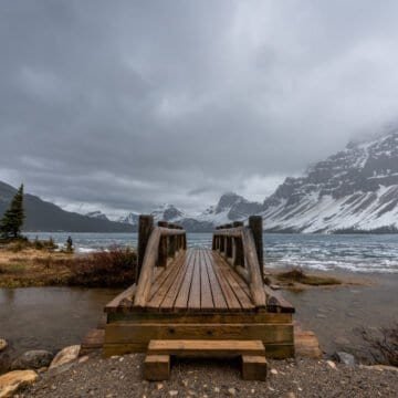 Wooden bridge over small water body leading to partially frozen lake with snow-covered mountains.