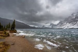 Partially frozen lake with snowy mountains and pine trees under cloudy sky.