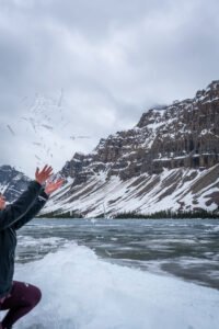 Person in dark jacket throws snow by frozen lake with snow-covered mountains under cloudy sky