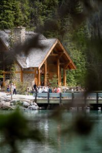 People gathered near a stone and wood lodge by a lake with evergreen trees and a bridge.