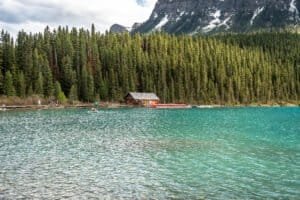 Turquoise lake bordered by evergreen forest, a wooden cabin, red canoes, and snow-capped mountains.