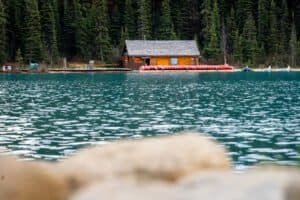 Wooden cabin by turquoise lake with red canoes on dock and evergreen forest background.