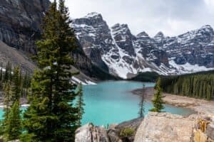 Turquoise lake surrounded by evergreen forest, rocky terrain, and snow-capped mountains.
