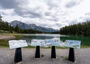 Mountain lake with four informational signboards on the shore and forested mountains under cloudy sky