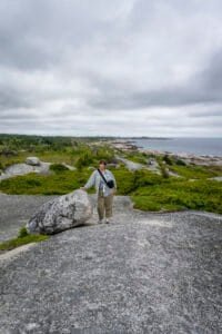 Person standing on rocky coastal terrain with green shrubs and ocean under a cloudy sky.