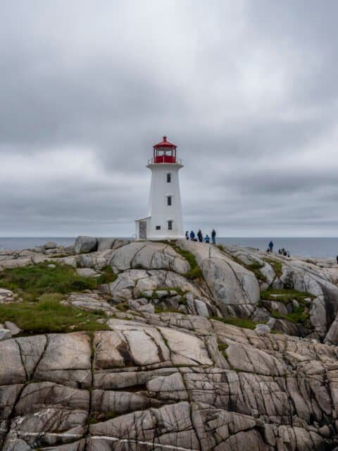 White lighthouse with red top on rocky coast overlooking ocean under cloudy sky with people nearby